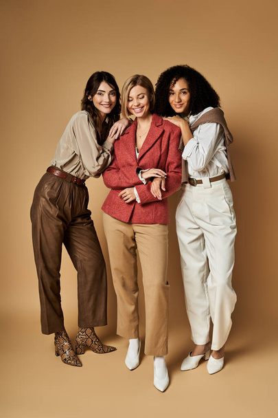 Three young, beautiful women of diverse cultures and backgrounds stand together in stylish attire on a beige background. - Photo, Image