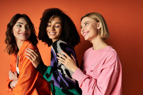Νεαρές πολυπολιτισμικές γυναίκες με ζωντανά ρούχα στέκονται μαζί σε έναν πορτοκαλί τοίχο, αποπνέοντας εμπιστοσύνη και φιλία. - Φωτογραφία, εικόνα