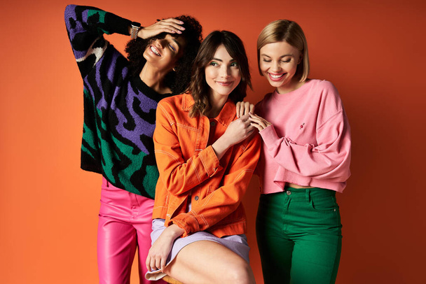 Tre giovani donne in abiti vivaci in posa su uno sfondo arancione, in mostra l'amicizia e la diversità culturale. - Foto, immagini
