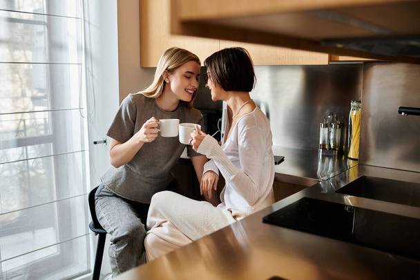 Mladý lesbický pár svázaný nad šálky kávy, sedí blízko a chatuje v útulné kuchyni uvnitř hotelového pokoje. - Fotografie, Obrázek