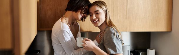 A young lesbian couple standing together in a hotel kitchen, holding coffee and engaging in conversation. - Foto, Bild