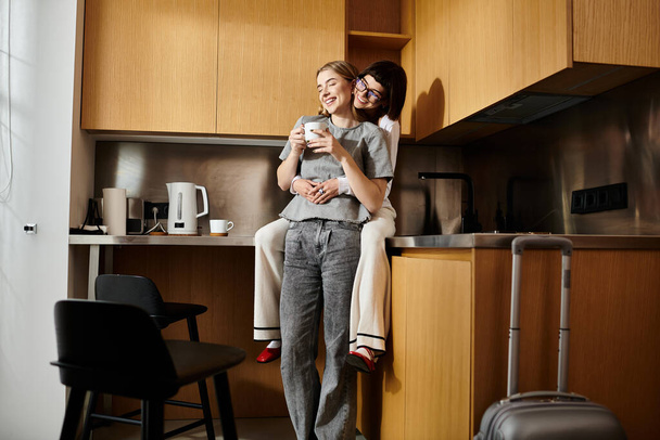 Mladý lesbický pár stojí v kuchyni hotelového pokoje, každý z nich drží šálek kávy, jak začínají svůj den. - Fotografie, Obrázek