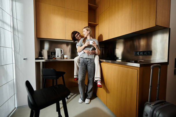 Ένας άντρας κρατά τρυφερά μια γυναίκα σε μια ζεστή κουζίνα, απολαμβάνοντας και οι δύο φλιτζάνια καφέ σε ένα δωμάτιο ξενοδοχείου ρύθμιση. - Φωτογραφία, εικόνα