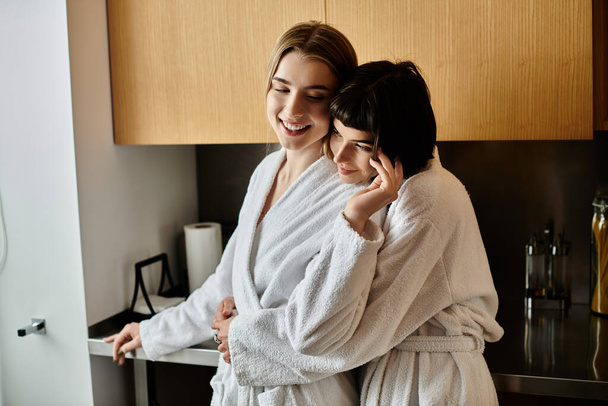 Een jong lesbisch paar in badjassen staan samen, het delen van een teder moment in een gezellige keuken setting. - Foto, afbeelding