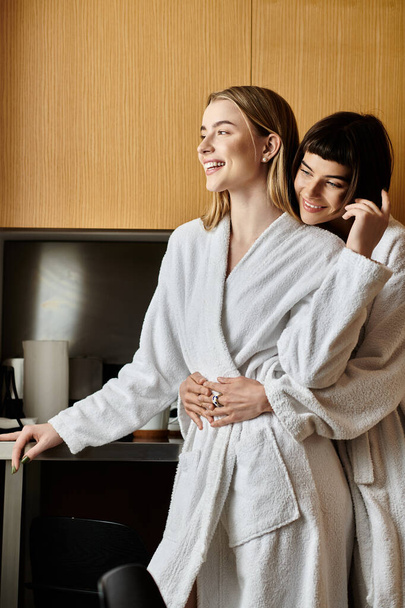 Two young women in bath robes standing close, exuding love and connection in a cozy hotel room setting. - Фото, изображение