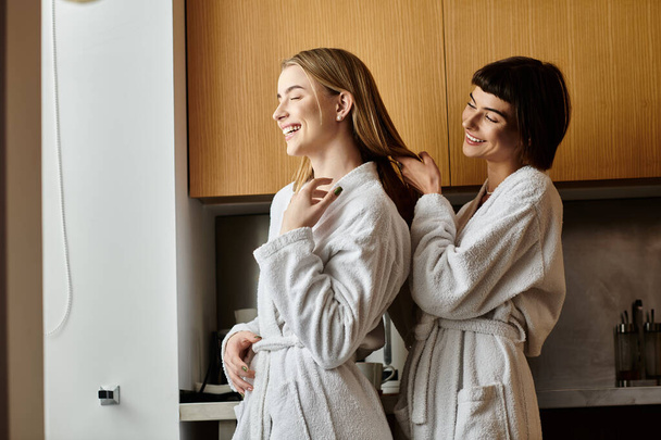 A young lesbian couple standing in a cozy kitchen wearing bath robes. - Zdjęcie, obraz