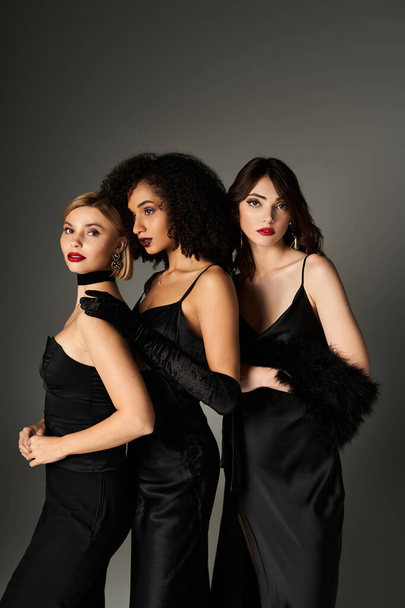 Drie jonge, mooie interraciale vrouwen staan verenigd en presenteren diversiteit in elegante zwarte jurken, tegen een grijze achtergrond. - Foto, afbeelding