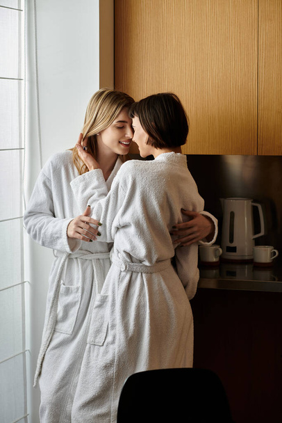 Two young women, a lesbian couple, stand close in bath robes inside a cozy hotel room, sharing a tender moment. - Foto, immagini