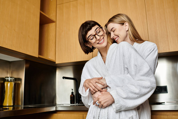Ein junges lesbisches Paar in Bademänteln teilt einen freudigen Moment, während es zusammen in einer warmen und einladenden Küche steht. - Foto, Bild