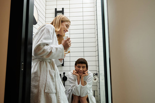 Μια γυναίκα με μπουρνούζι βουρτσίζει τα δόντια της ενώ ο συνεργάτης της βλέπει στο μπάνιο του ξενοδοχείου.. - Φωτογραφία, εικόνα