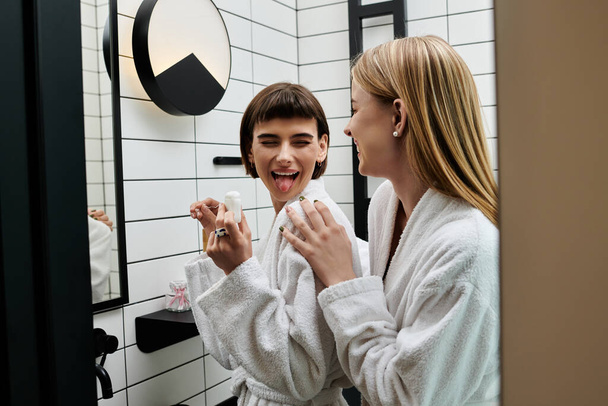 Μια νεαρή γυναίκα με μπουρνούζι να πλένει τα δόντια της μπροστά από έναν καθρέφτη στο μπάνιο ενός ξενοδοχείου.. - Φωτογραφία, εικόνα
