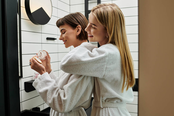 Twee jonge vrouwen in witte gewaden staren naar hun weerspiegeling in een spiegel in een hotelbadkamer. - Foto, afbeelding