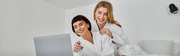 Young lesbian couple in white robes exploring something intriguing on a laptop together. - Foto, immagini