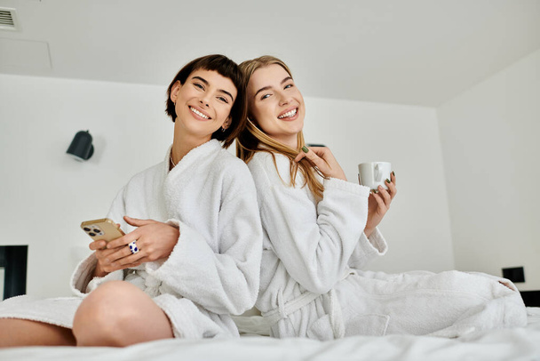 Ένα εκπληκτικό λεσβιακό ζευγάρι με μπουρνούζια απολαμβάνει μια στιγμή χαλάρωσης ενώ κάθεται στην κορυφή ενός πολυτελούς κρεβατιού μέσα σε ένα δωμάτιο ξενοδοχείου. - Φωτογραφία, εικόνα