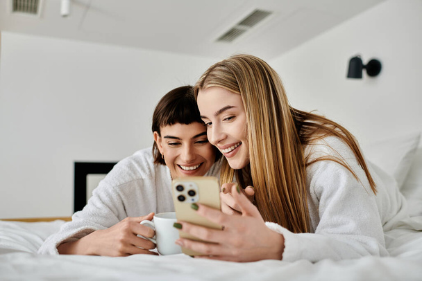 Una bella coppia lesbica in accappatoi giaceva a letto, profondamente assorto nel morbido bagliore di uno schermo del telefono cellulare. - Foto, immagini