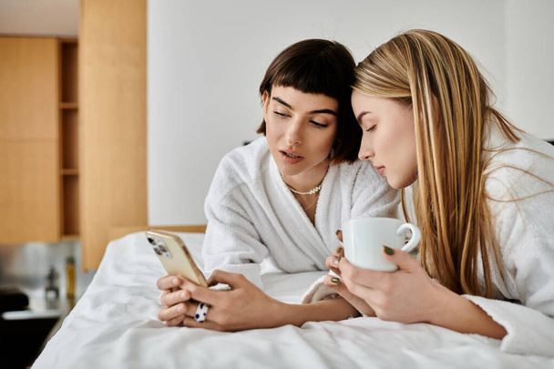 A beautiful lesbian couple in bath robes lay on bed, engrossed in cell phone - Photo, Image