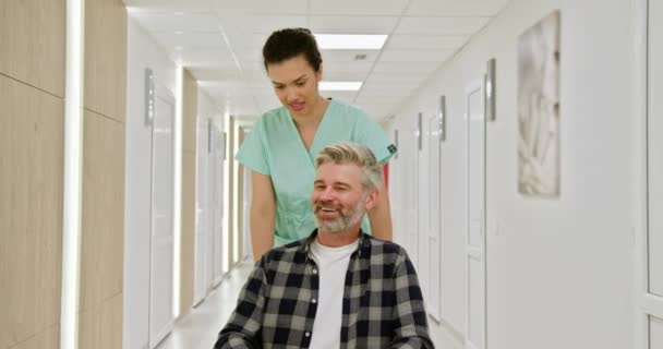 Медсестра помогает пожилому человеку в инвалидном кресле через больничный коридор. Медсестра помогает пожилому человеку в инвалидном кресле по ярко освещенному коридору больницы, делая акцент на уходе и медицинской поддержке. - Кадры, видео