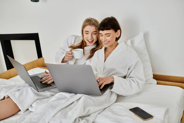 Dos mujeres vestidas con túnicas blancas colaboran en un portátil mientras descansan en una cómoda cama dentro de una habitación de hotel. - Foto, imagen