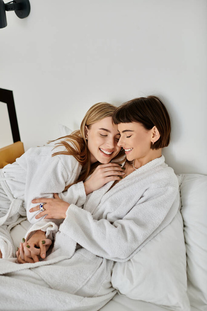 Ένα όμορφο λεσβιακό ζευγάρι με μπουρνούζια χαλαρώνει σε ένα βελούδινο κρεβάτι, αγκαλιασμένο κάτω από μια ζεστή κουβέρτα.. - Φωτογραφία, εικόνα