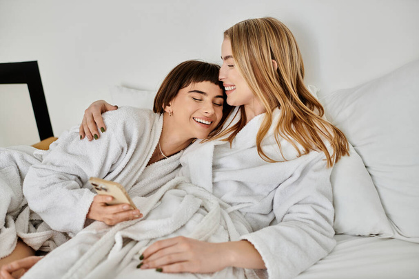A lesbian couple in bath robes relax, laying in bed under a cozy blanket in a hotel room. - Foto, Imagem