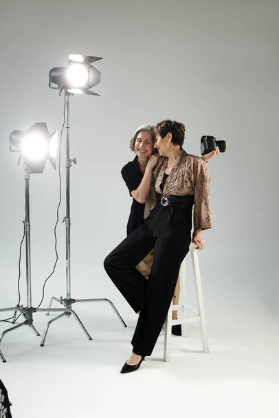 Μεσήλικες λεσβίες ζευγάρι, μία ως φωτογράφος με κάμερα, άλλη ως μοντέλο, κάθεται σε σκαμνί στο φωτογραφείο. - Φωτογραφία, εικόνα