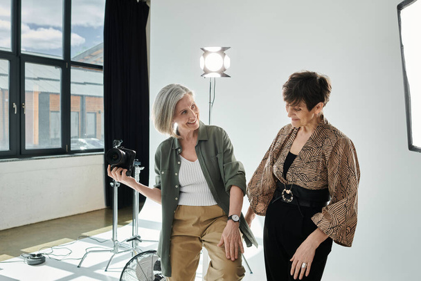 Para lesbijek w średnim wieku w studiu fotograficznym, jedna trzymająca kamerę, a druga pozująca w studiu - Zdjęcie, obraz
