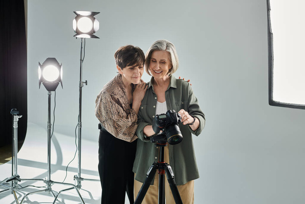 Lesbický pár středního věku ve fotostudiu, jeden s kamerou a druhý jako modelka, stojící vedle sebe. - Fotografie, Obrázek