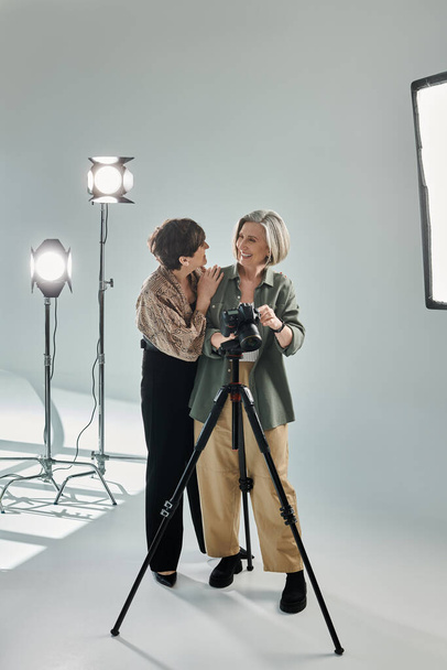 Een lesbisch stel van middelbare leeftijd in een fotostudio; de een achter de camera, de ander knuffelt haar - Foto, afbeelding