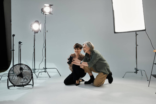 Een fotograaf en model, een lesbisch stel van middelbare leeftijd, knielen voor de camera in een fotostudio. - Foto, afbeelding