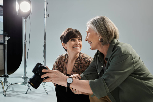 Para lesbijek w średnim wieku w studiu fotograficznym; jedna kobieta z aparatem fotograficznym, a druga z uśmiechem - Zdjęcie, obraz