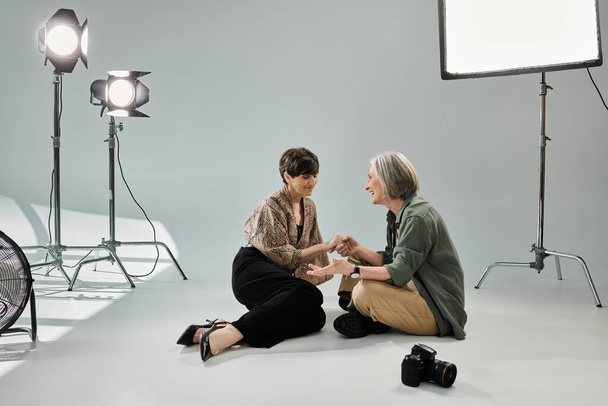 Keski-ikäinen lesbo pari valokuvastudiossa, yksi valokuvaaja kameran kanssa, toinen malli, istuu maassa. - Valokuva, kuva
