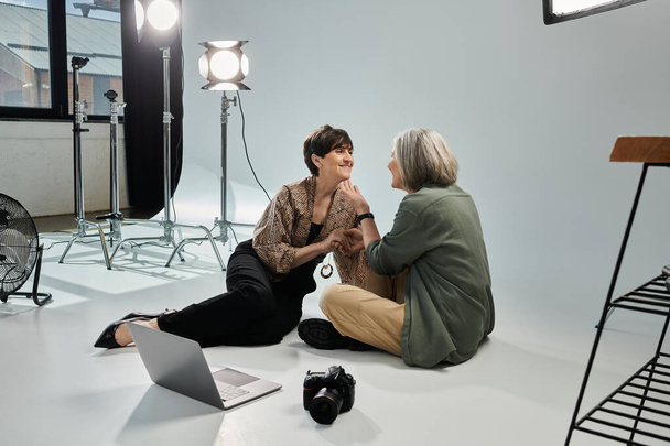 Una pareja de lesbianas de mediana edad, una como fotógrafa y la otra como modelo, sentadas en un estudio fotográfico. - Foto, imagen