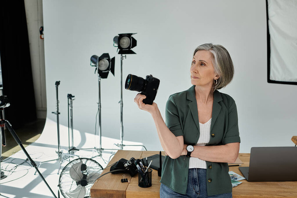 Een vrouw van middelbare leeftijd houdt een camera vakkundig vast in een professionele studio-omgeving, gefocust en klaar om de perfecte opname te maken. - Foto, afbeelding