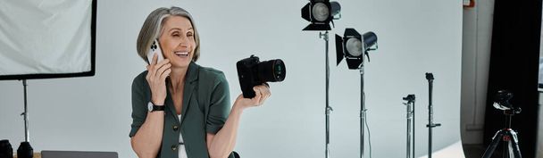 Una mujer de mediana edad sosteniendo la cámara y hablando en un teléfono inteligente en un vibrante estudio fotográfico con equipo profesional e iluminación. - Foto, imagen