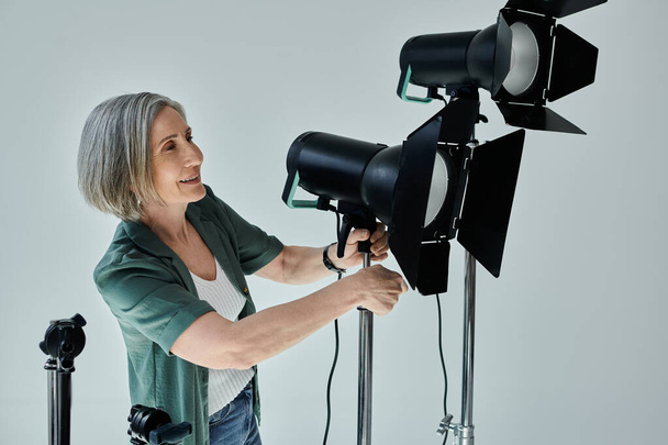 Kobieta w średnim wieku trzyma światło podczas ustawiania statywu w profesjonalnym studiu fotograficznym - Zdjęcie, obraz