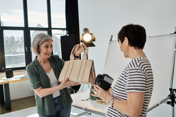 Μια μεσήλικη γυναίκα που κρατάει μια τσάντα και μια κάμερα σε ένα μοντέρνο φωτογραφικό στούντιο. - Φωτογραφία, εικόνα