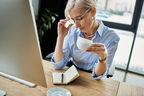 A middle aged businesswoman with short hair enjoys a cup of coffee while working in her office - Photo, Image