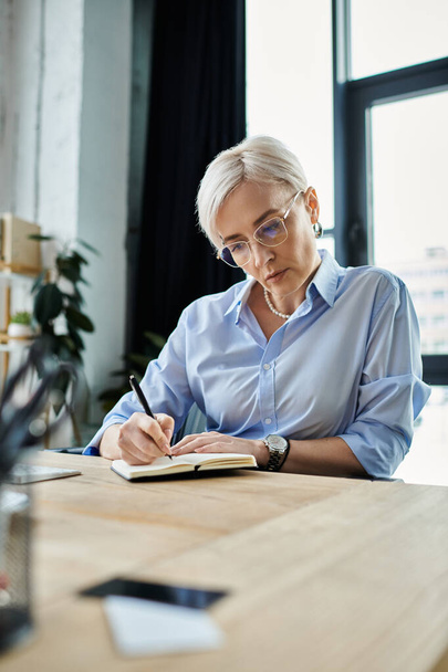 A middle-aged businesswoman with short hair sits at a table writing in a book in her office. - Photo, Image