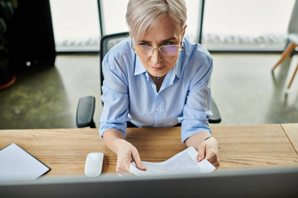 Orta yaşlı, kısa saçlı bir iş kadını menopoz sırasında ofis masasındaki bir kağıt parçasına odaklanıyor.. - Fotoğraf, Görsel