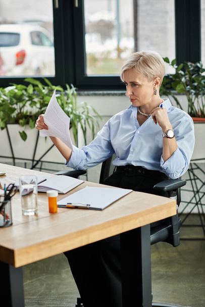 Una donna d'affari di mezza età concentrata con i capelli corti seduta con documento alla scrivania in un ambiente di ufficio, sensazione di caldo durante la menopausa - Foto, immagini