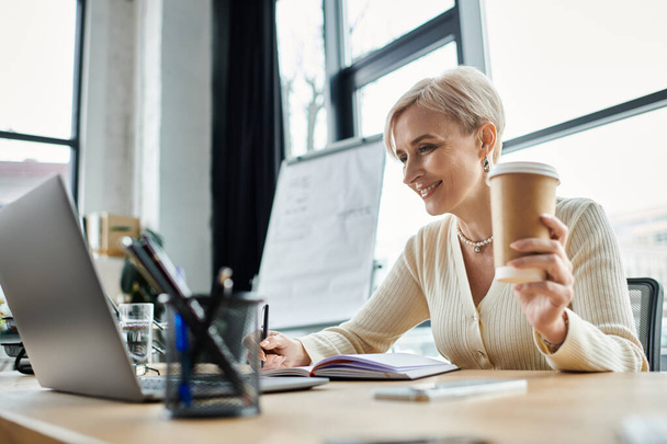 Una mujer de negocios de mediana edad con el pelo corto trabajando diligentemente en su computadora portátil mientras disfruta de una taza de café en su escritorio. - Foto, Imagen