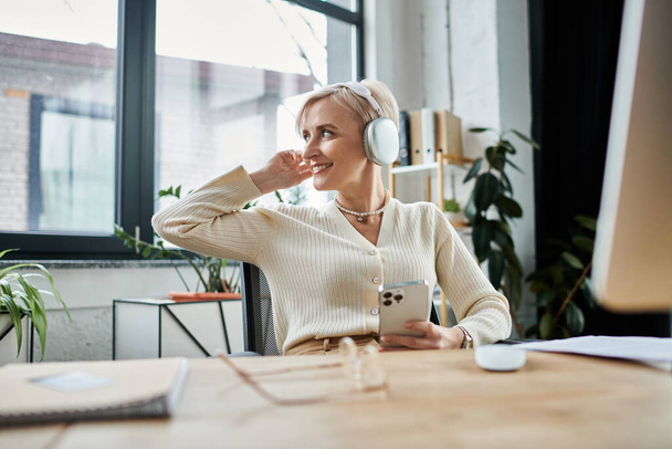 Una donna d'affari di mezza età con i capelli corti seduta alla scrivania, che utilizza attivamente un telefono cellulare per il lavoro. - Foto, immagini