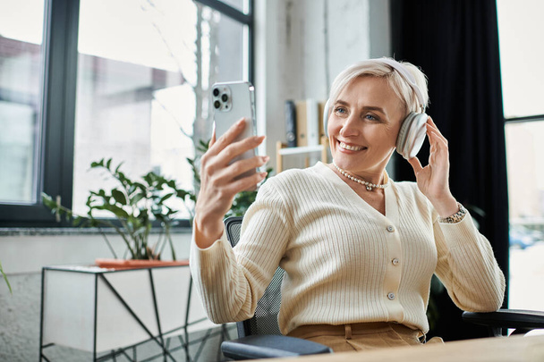 Μια μεσήλικη επιχειρηματίας με κοντά μαλλιά κάθεται σε ένα γραφείο, μιλώντας στο κινητό της σε ένα μοντέρνο περιβάλλον γραφείου.. - Φωτογραφία, εικόνα