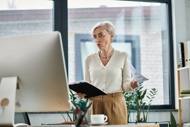 Geschäftsfrau mittleren Alters mit kurzen Haaren steht im Büro und hält Papier vor den Computer. - Foto, Bild