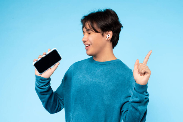 Asian young guy in a blue sweater is happily listening to music on wireless earbuds while holding his smartphone. He points upwards with a smile on his face - Фото, изображение