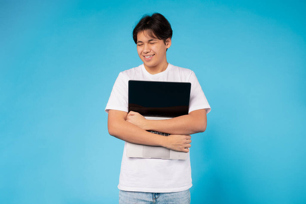 Aziatische jongeman met een wit t-shirt en jeans staat tegen een levendige blauwe achtergrond. Hij glimlacht en lijkt tevreden terwijl hij een laptop dicht bij zijn borst houdt. - Foto, afbeelding
