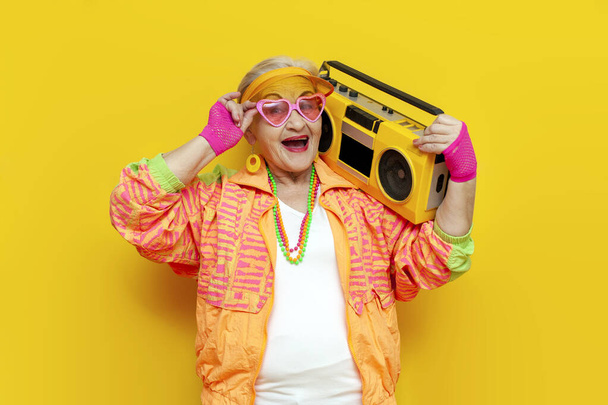 funny crazy granny in hipster clothes listening to music on tape recorder and singing on yellow isolated background, elderly cool woman dancing at party and screaming - Photo, Image
