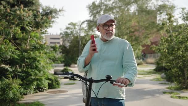 Abuelo latino con bicicleta en una llamada telefónica hablando con la familia en el parque público - Metraje, vídeo