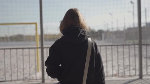 Young Woman Standing by a Sports Field on a Sunny Day. A young woman in casual attire with a backpack stands by a sports field, facing away, contemplating under a sunny sky. - Footage, Video