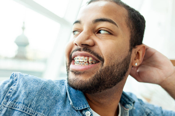 щасливий афроамериканський чоловік з брекетами сміється і дивиться у вікно в білому кафе, розслаблений чоловік в джинсовій сорочці мріє і посміхається - Фото, зображення